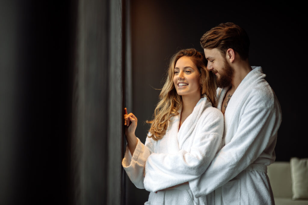A couple in white bathrobes standing together by a window, smiling