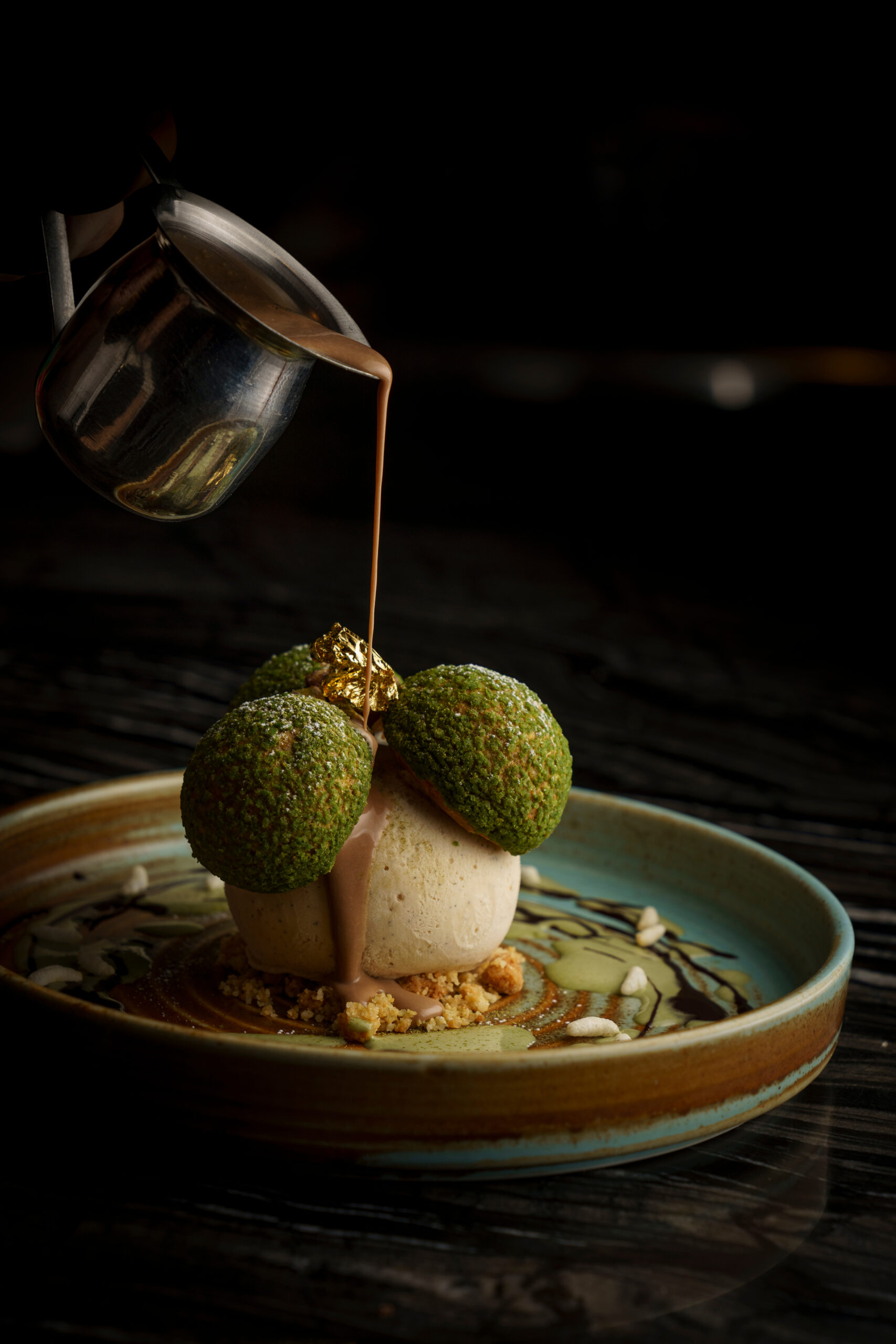 Sauce poured from a small jug over a plated dessert with moss green spheres, on a dark table